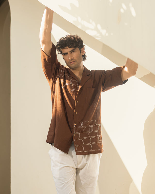 Man wearing a brown patterned shirt and white pants standing against a light background