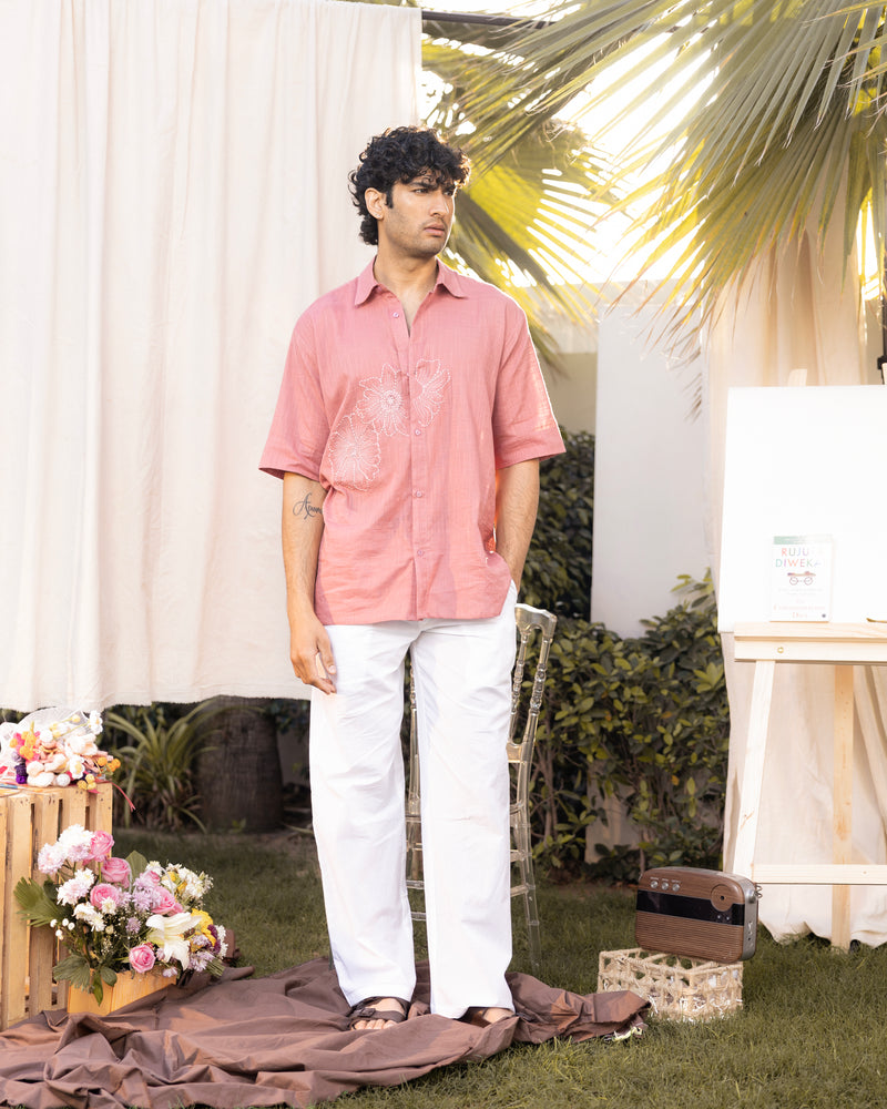 Man wearing a pink shirt and white pants standing outdoors with floral decorations and an easel in the background.