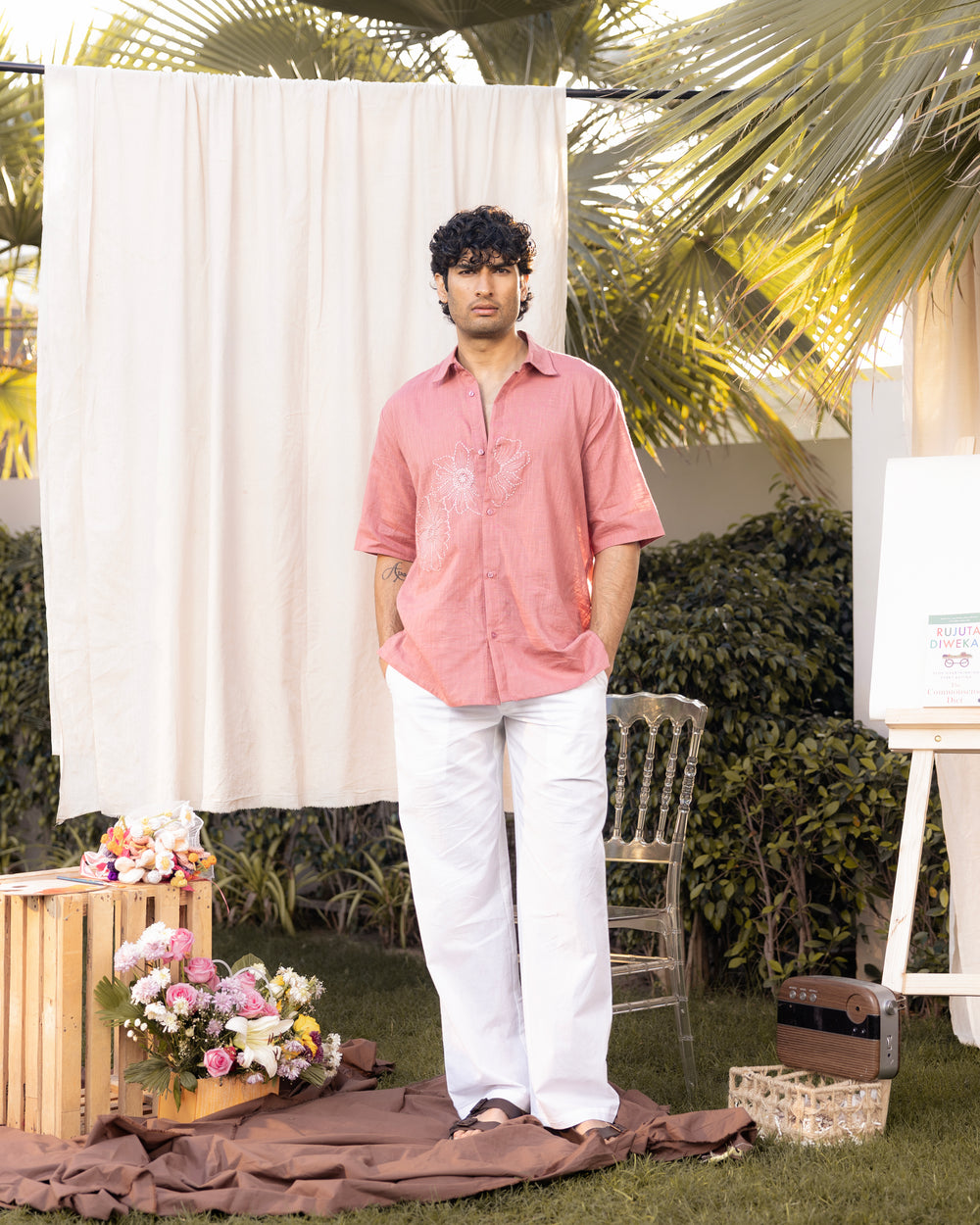 Man in a pink shirt and white pants standing outdoors with floral arrangements and a chair in the background.