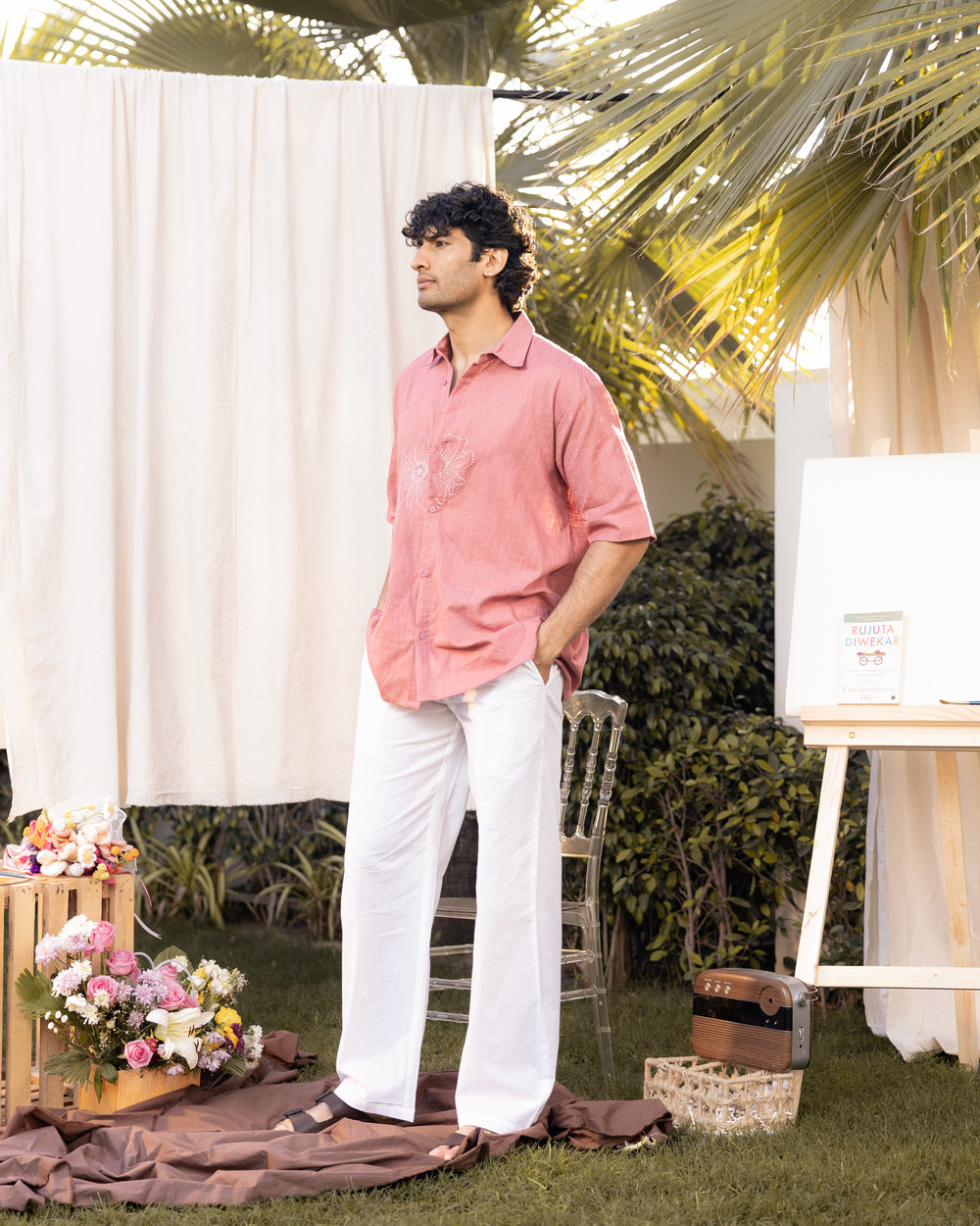 Man in a pink shirt and white pants standing outdoors with floral decorations and a chair.