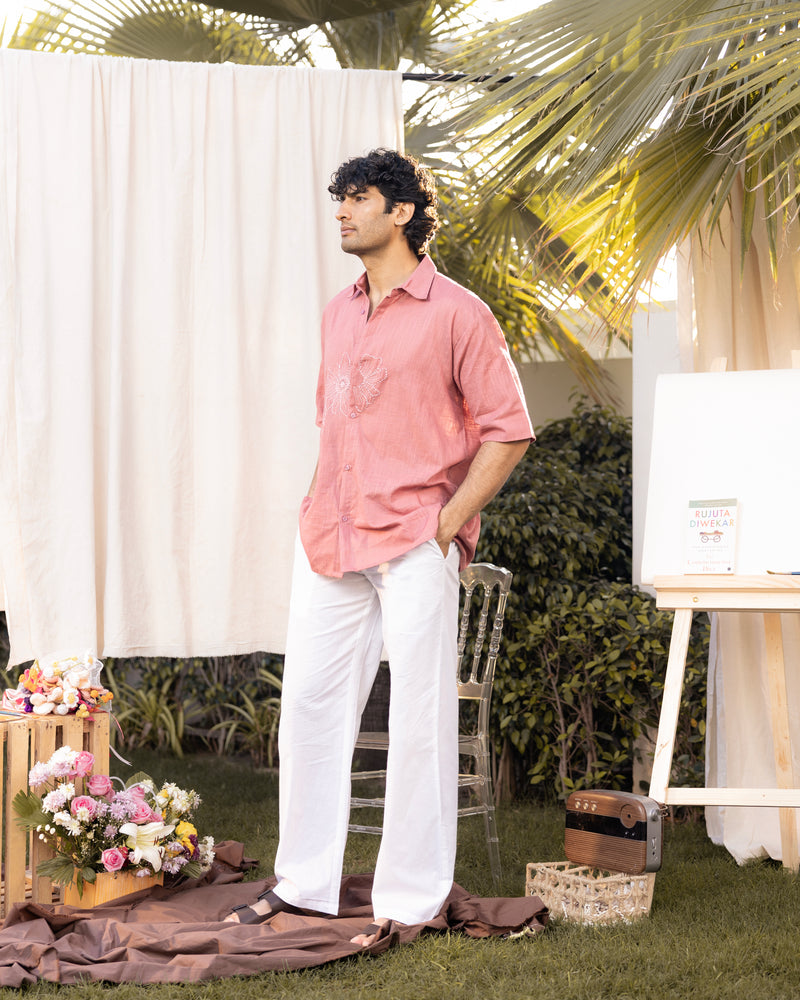 Man in a pink shirt and white pants standing outdoors with floral decorations and a chair.