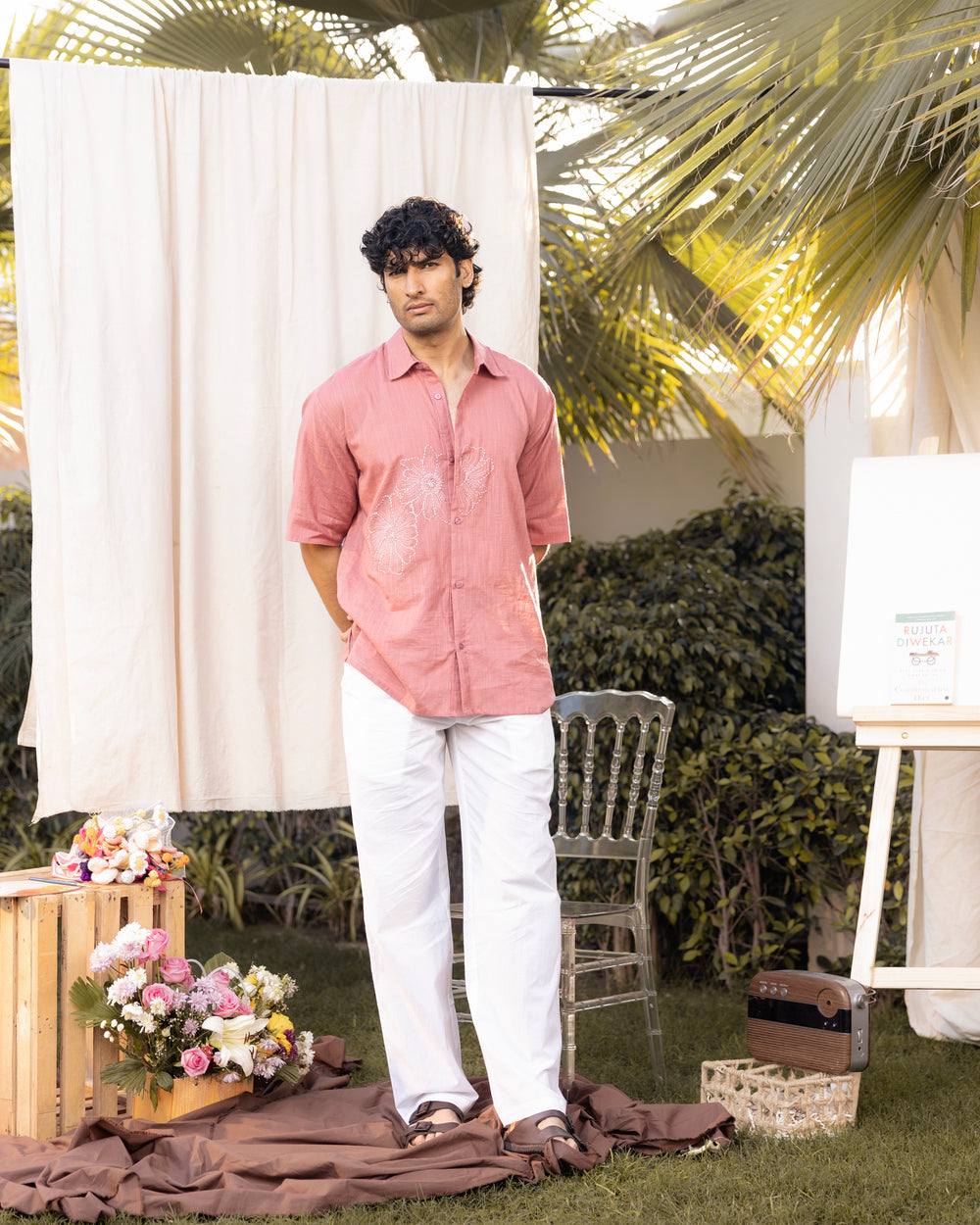 Man in a pink shirt and white pants standing outdoors with floral arrangements and a chair.