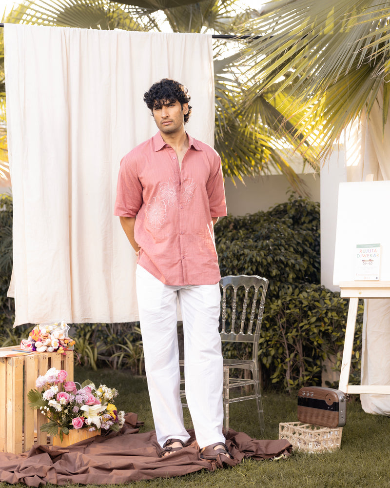 Man in a pink shirt and white pants standing outdoors with floral arrangements and a chair.