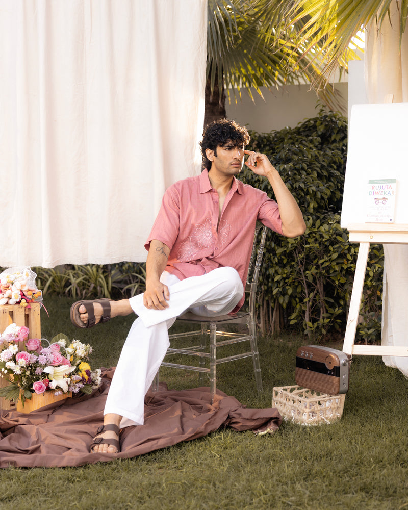 Man sitting outdoors in a pink shirt and white pants with plants and flowers around.