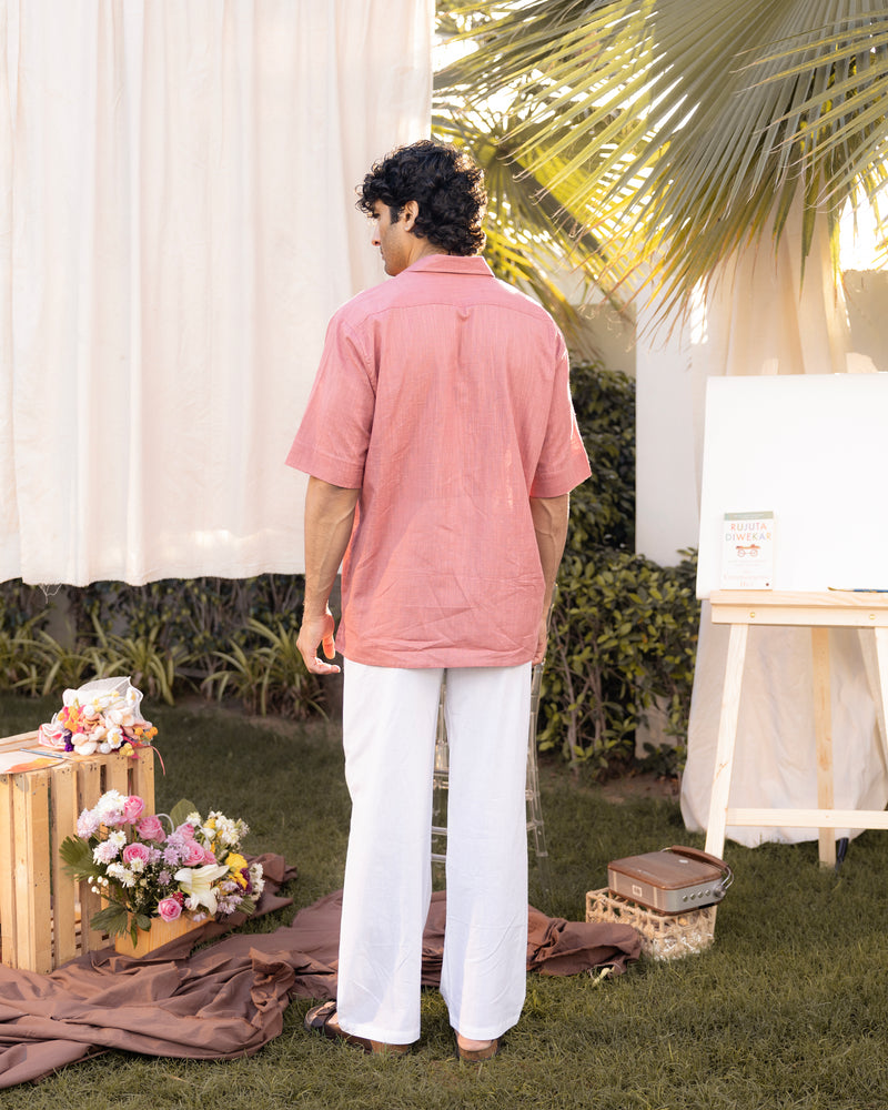 Person wearing a pink shirt and white pants standing outdoors with plants and decor in the background