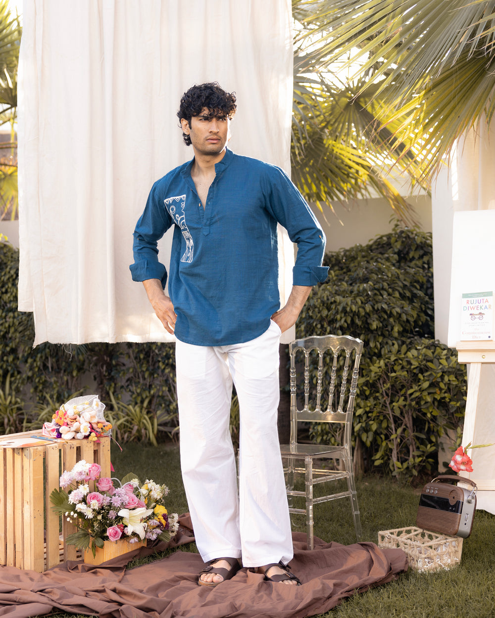 Man in a blue shirt and white pants standing outdoors with greenery and a chair in the background.
