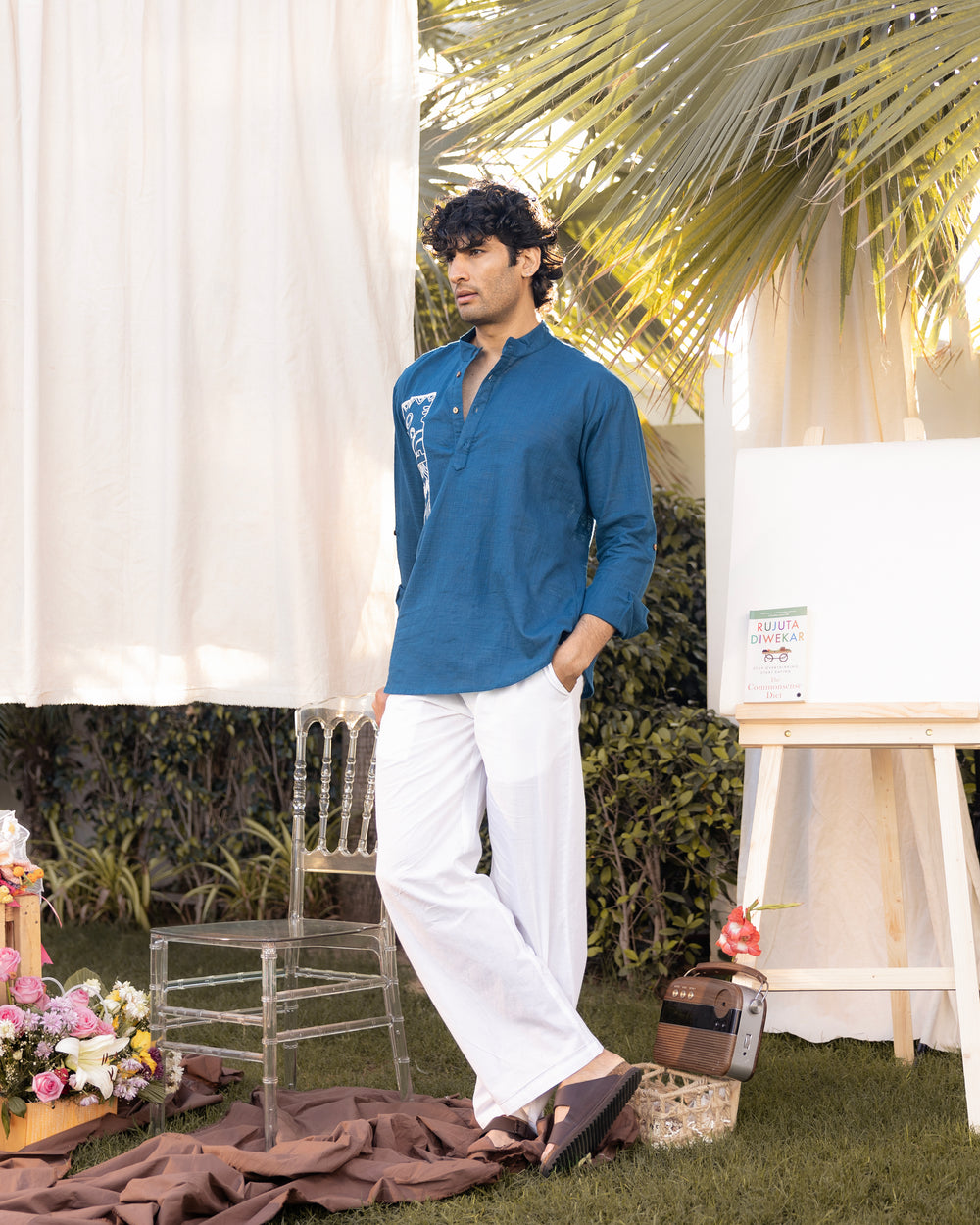 Man in a blue shirt and white pants standing outdoors with palm leaves and decor in the background
