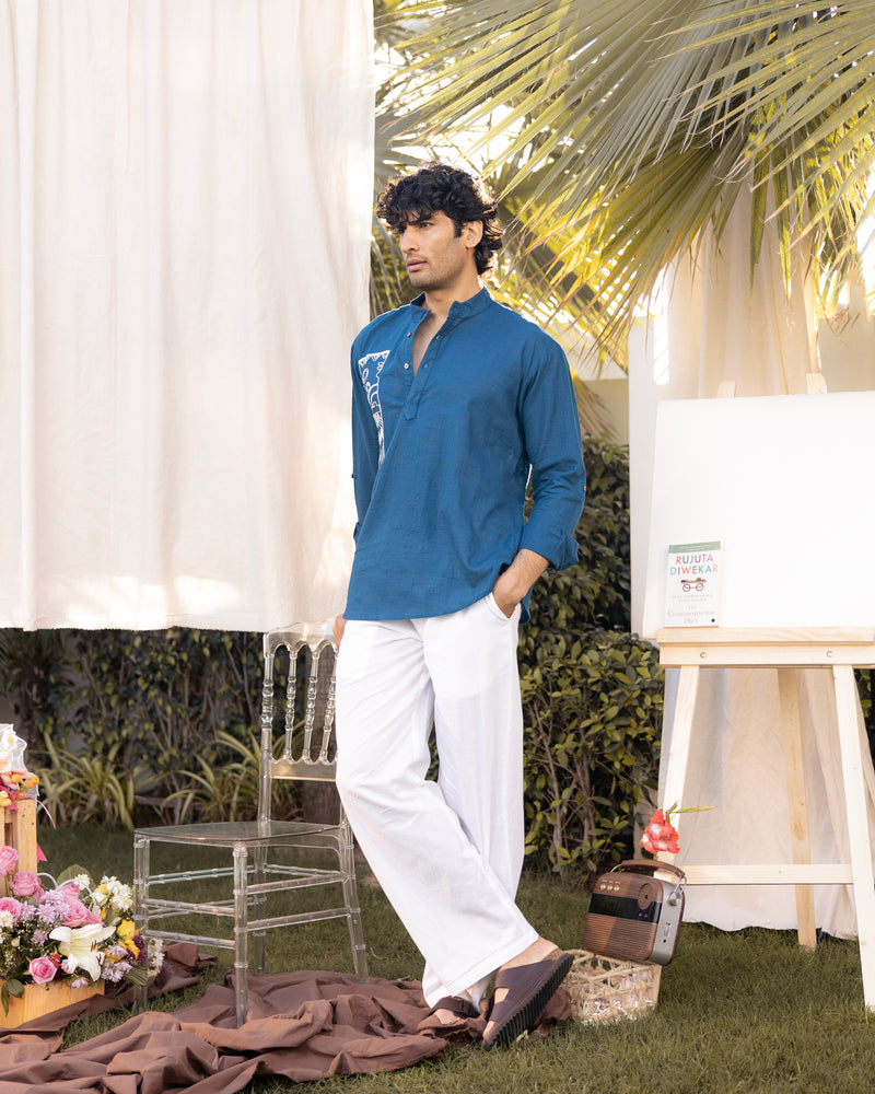 Man in a blue shirt and white pants standing outdoors with palm leaves and decor in the background