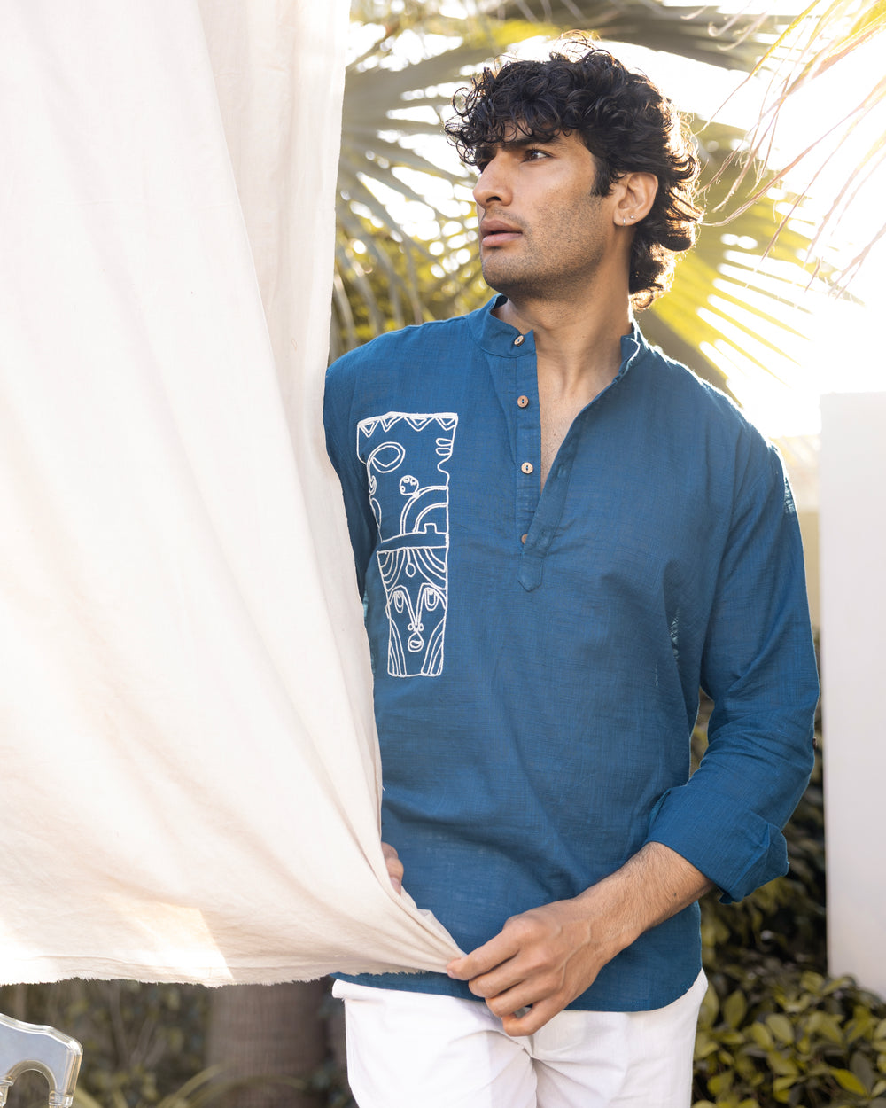 Man wearing a blue shirt with a white design, standing outdoors with greenery in the background.