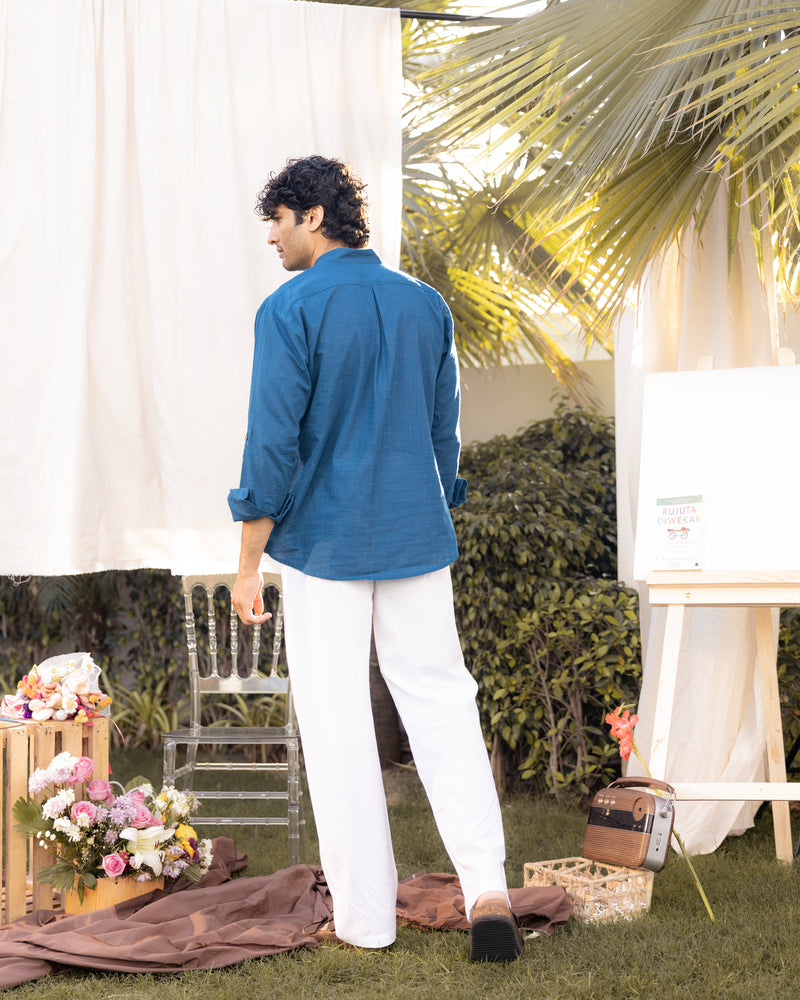 Man in a blue shirt and white pants standing outdoors with floral arrangements and a tent in the background.