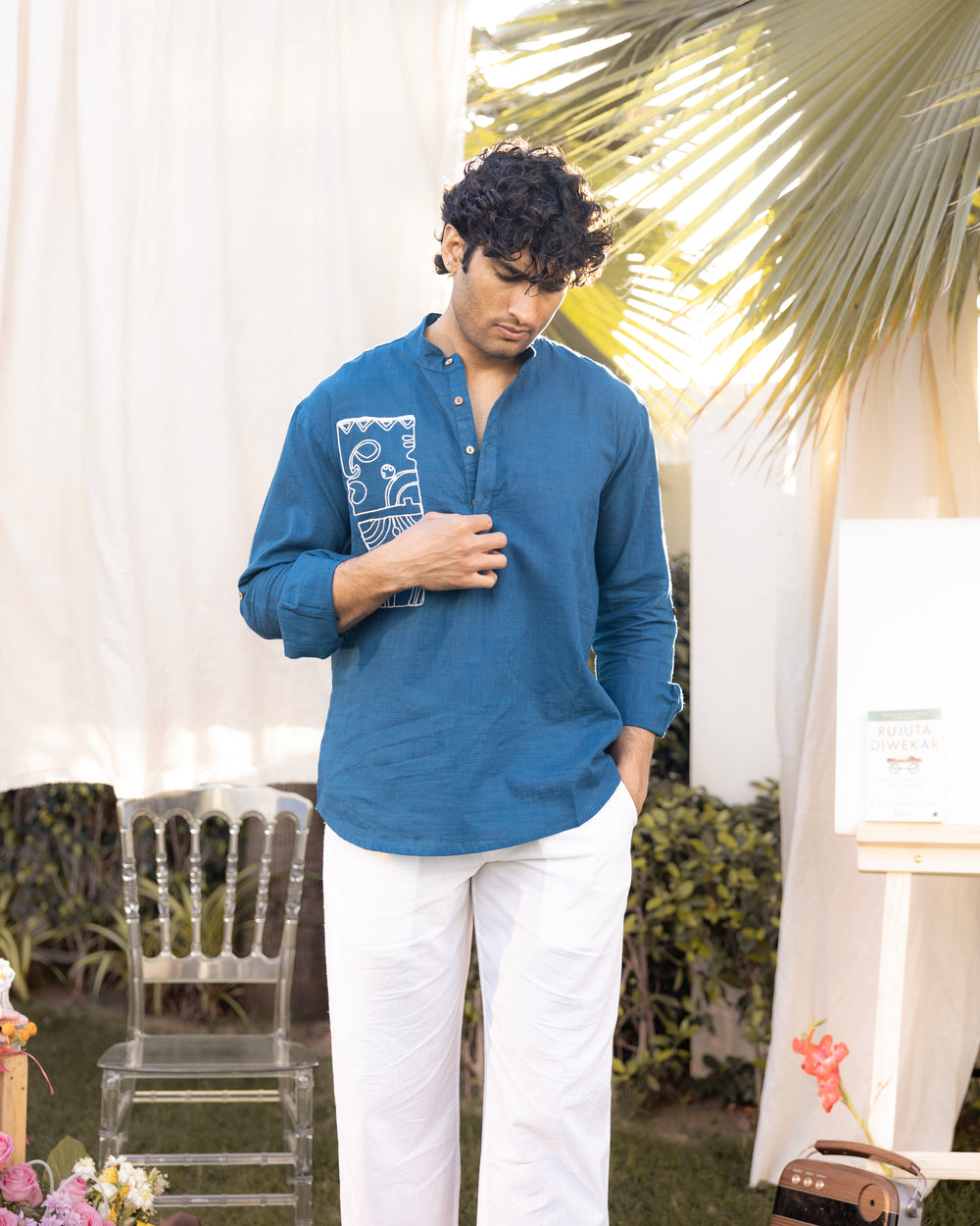Man wearing a blue shirt with white pants standing outdoors with plants and chairs in the background