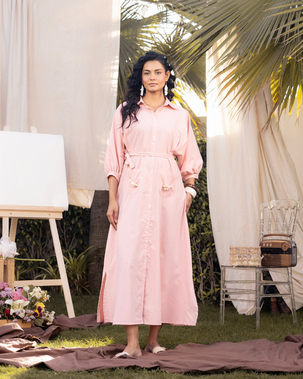 Woman in a pink dress standing outdoors with greenery and white curtains in the background