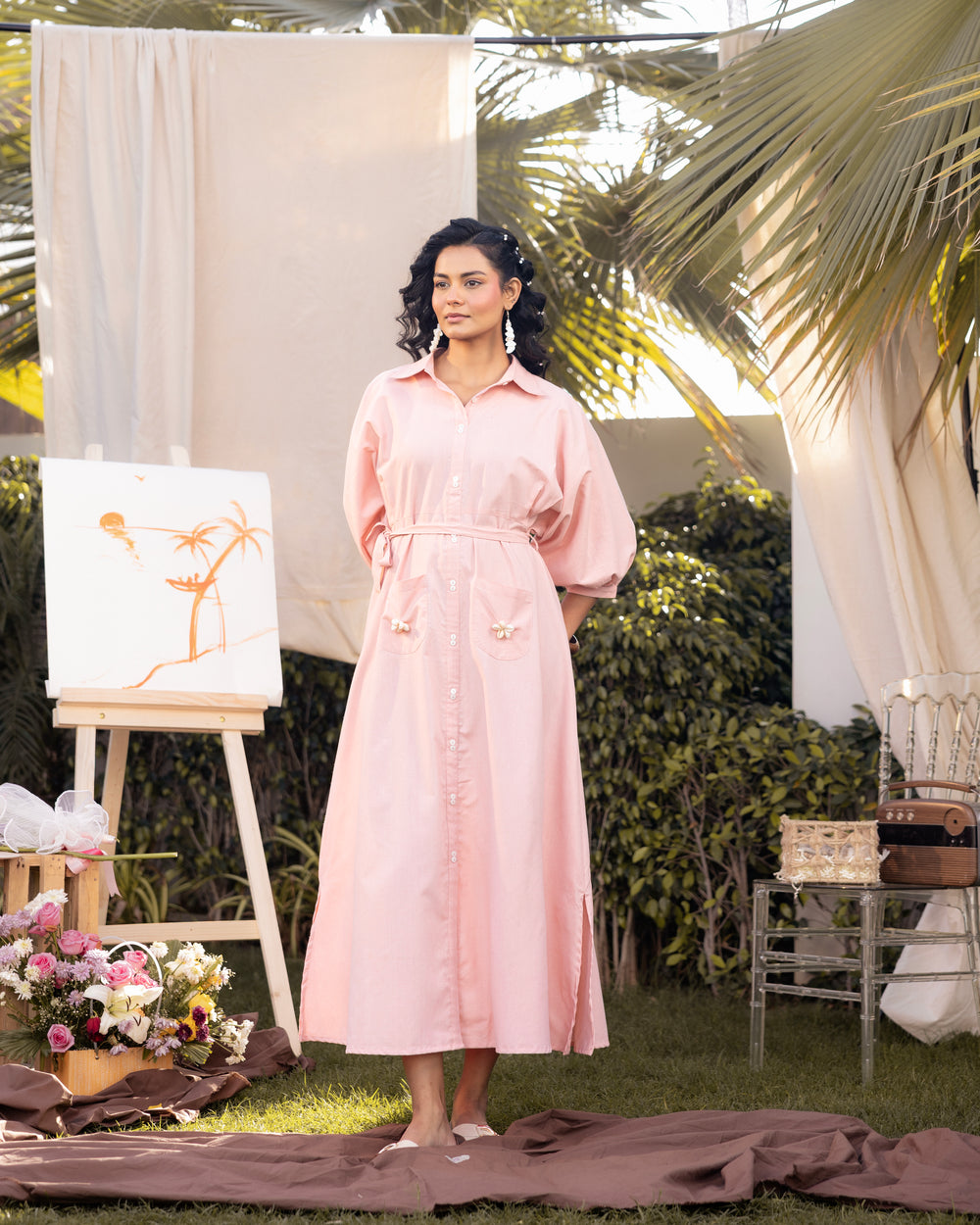 Woman in a pink dress standing outdoors with art supplies and plants around.