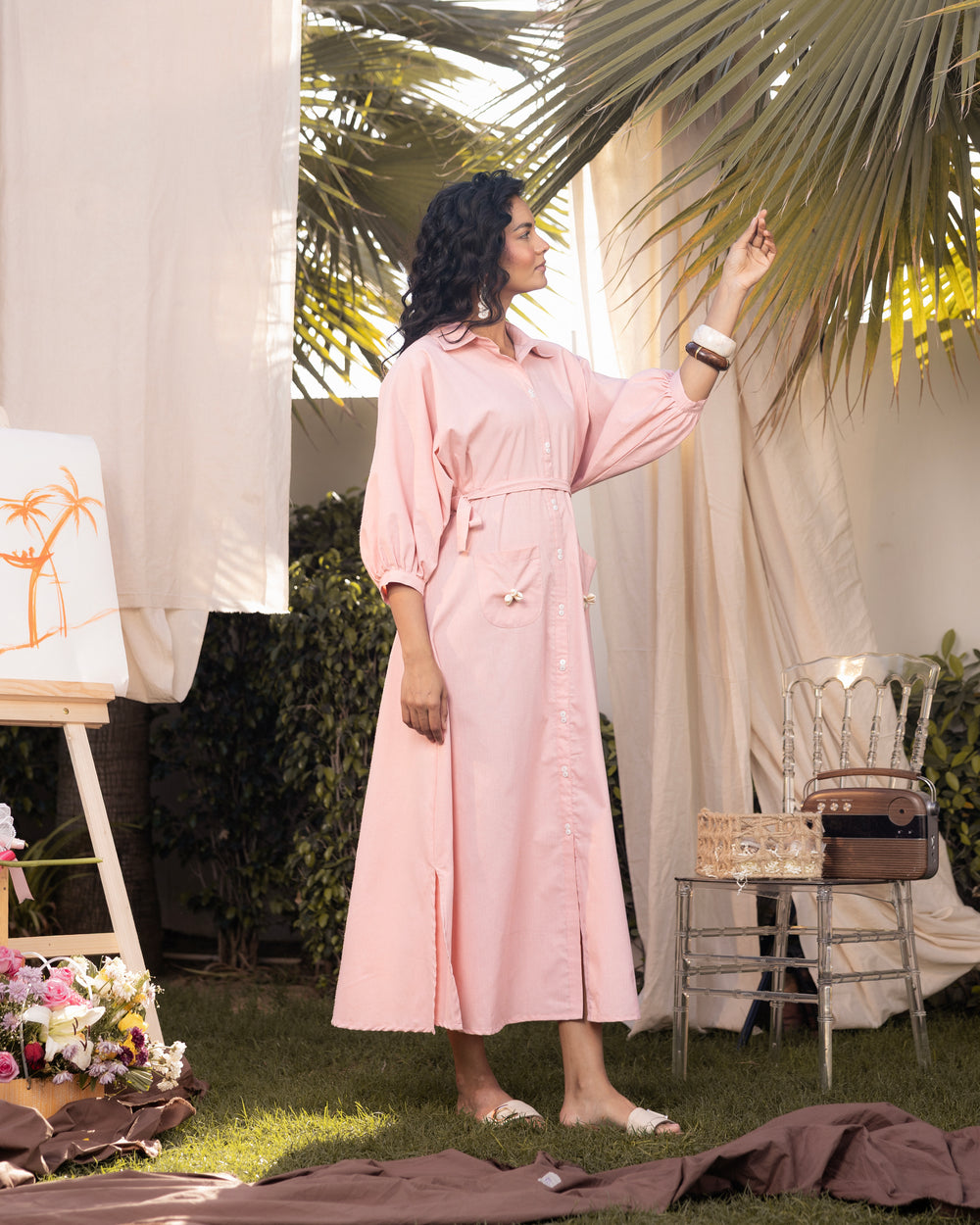 Woman in a pink dress standing outdoors with greenery and a chair in the background