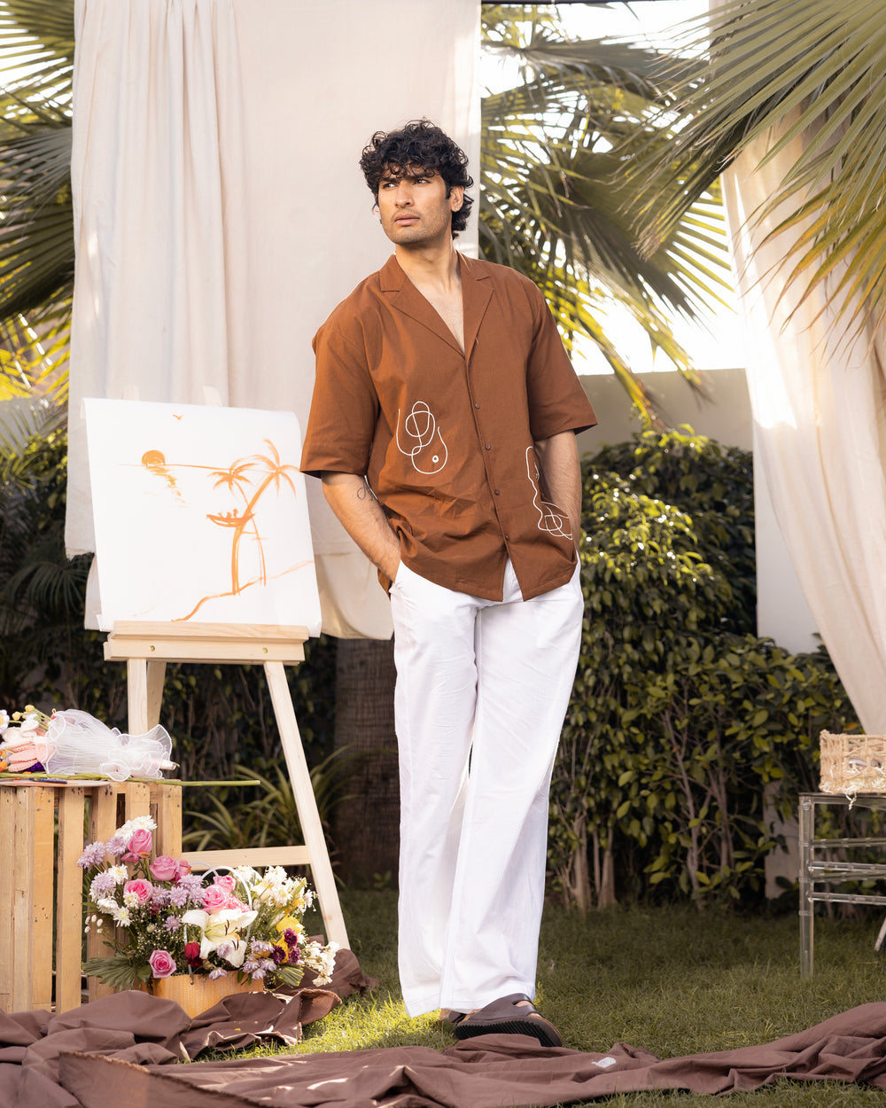 Man in brown shirt and white pants standing outdoors with art easel and flowers.