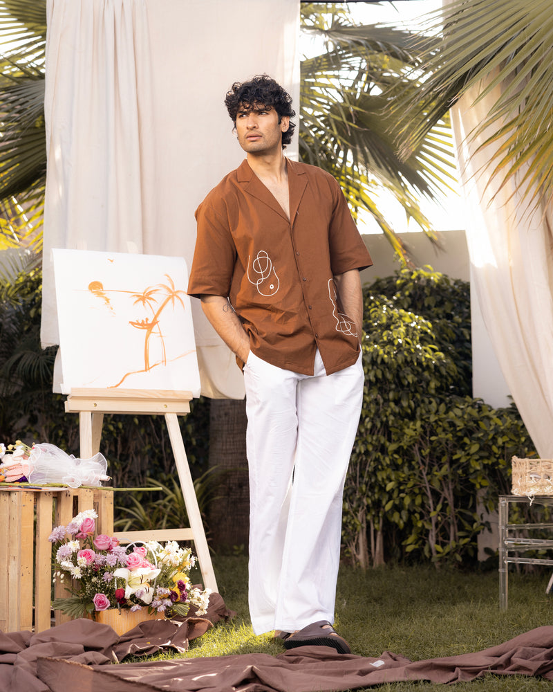 Man in brown shirt and white pants standing outdoors with art easel and flowers.