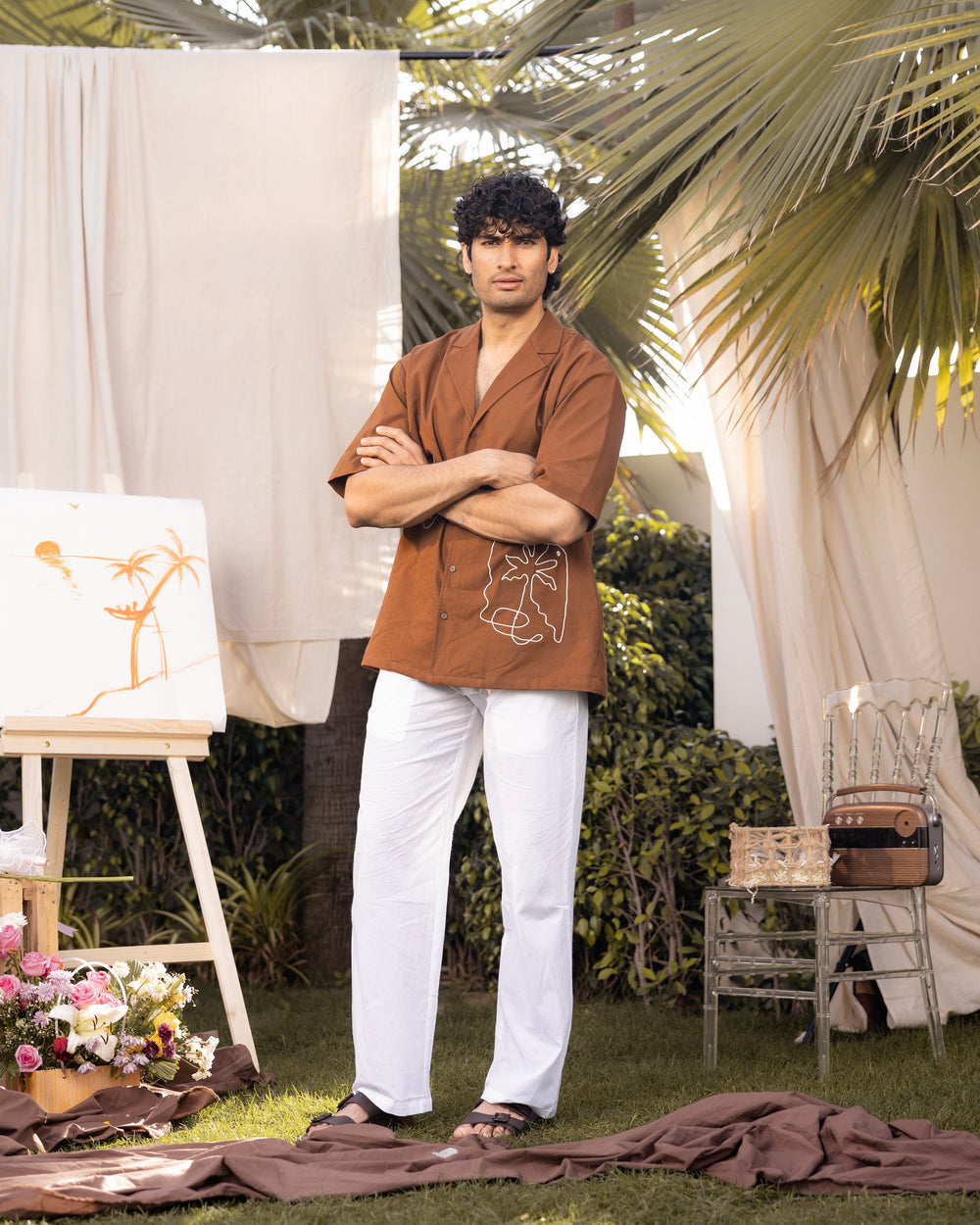 Man standing outdoors with arms crossed, wearing a brown shirt and white pants, with decorative elements in the background.
