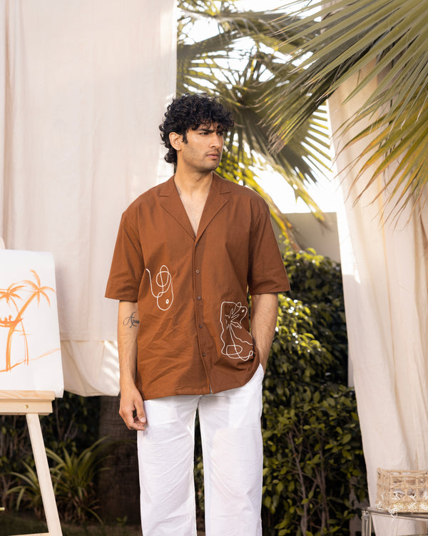 Man wearing a brown shirt with white pants standing outdoors with plants in the background
