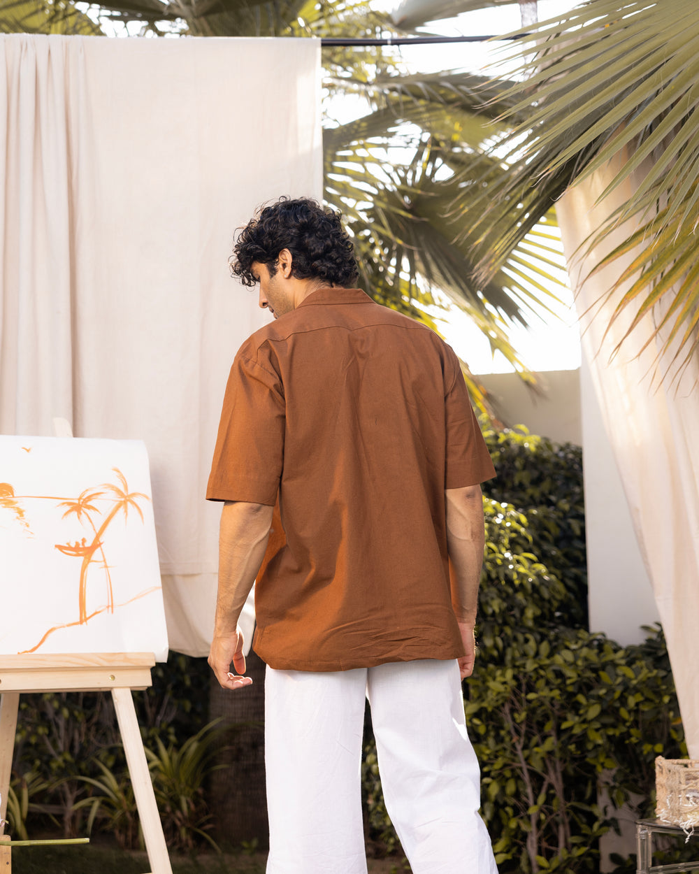 Man in a brown shirt and white pants standing outdoors with plants and a white curtain in the background.