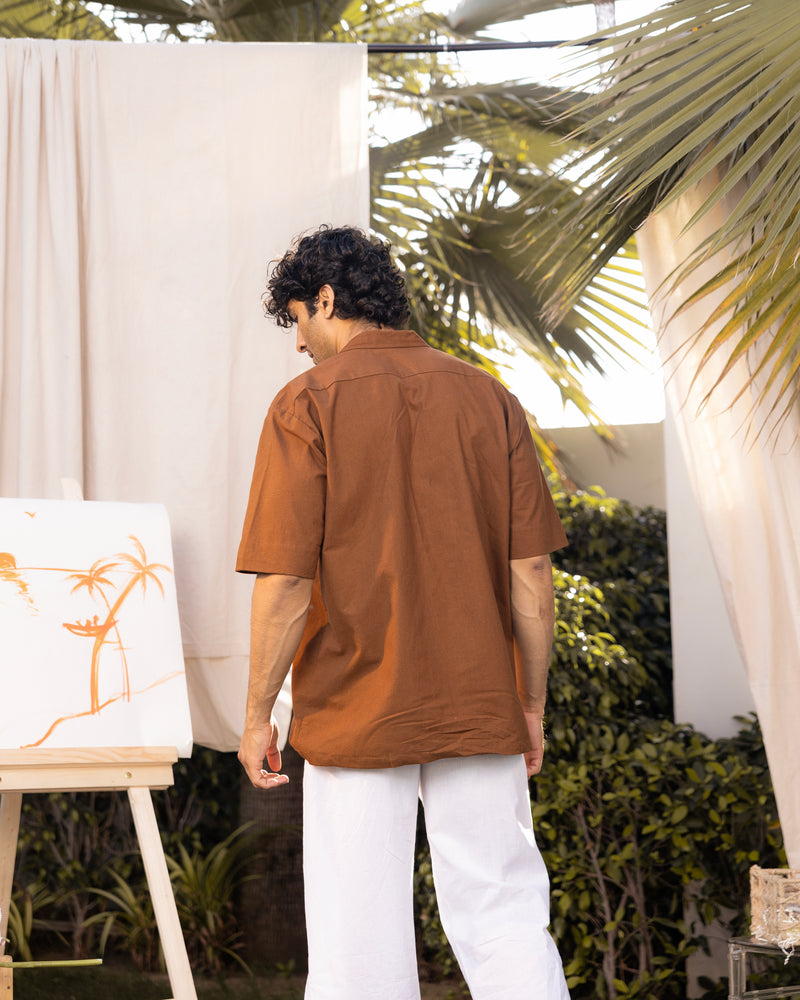 Man in a brown shirt and white pants standing outdoors with plants and a white curtain in the background.