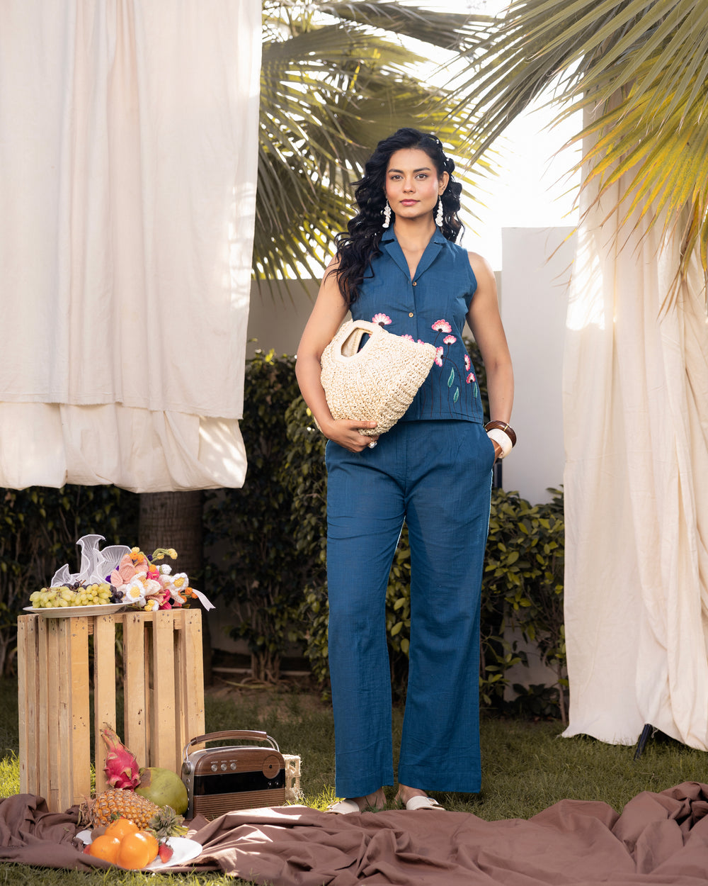 Woman in a blue outfit holding a woven bag outdoors with curtains and plants in the background