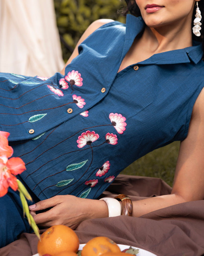 Woman wearing a blue dress with floral embroidery, sitting outdoors with flowers and oranges.
