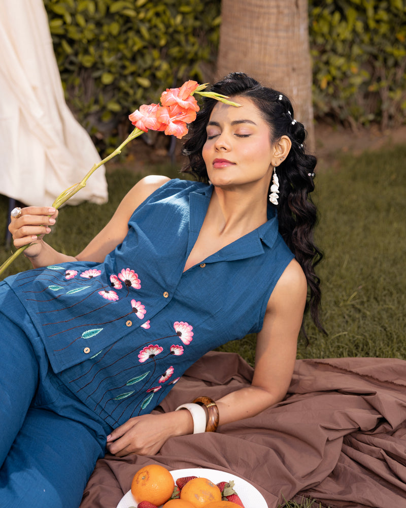 Woman in a blue embroidered outfit holding a pink flower, sitting outdoors with a brown blanket and fruit on a table.