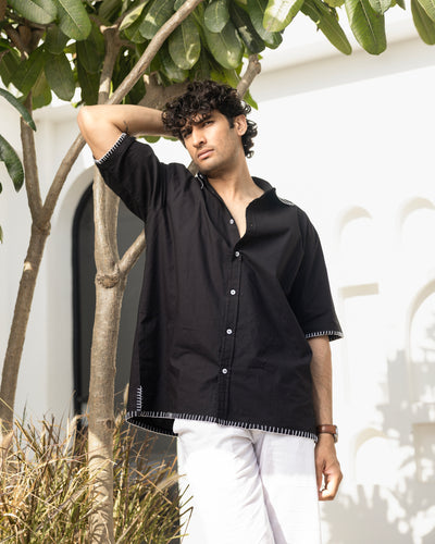 Man wearing a black shirt with white pants standing outdoors near a tree