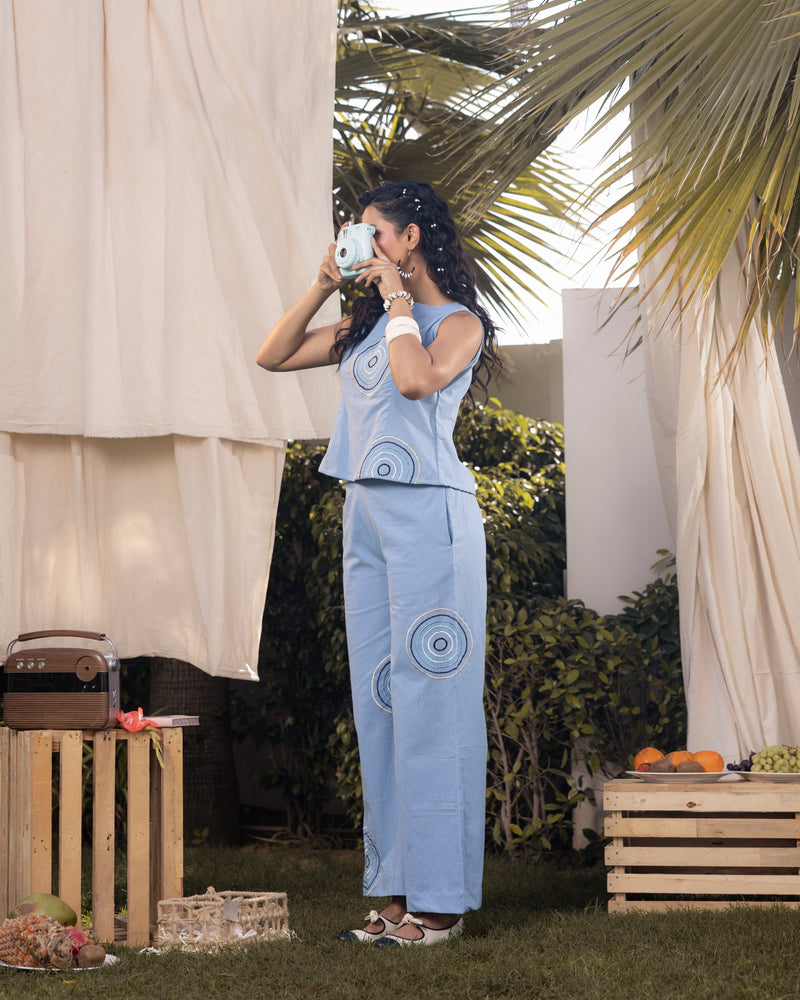 Woman in a light blue outfit drinking from a mug outdoors with plants and wooden crates in the background.