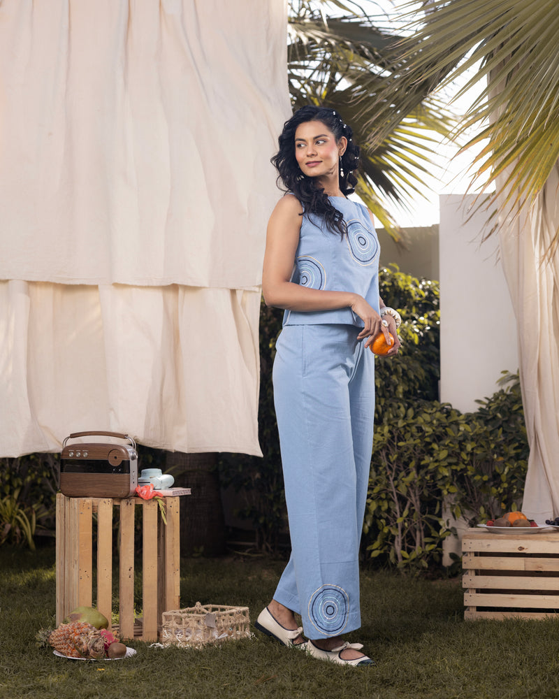 Woman in a light blue outfit standing outdoors with plants and a wooden crate in the background