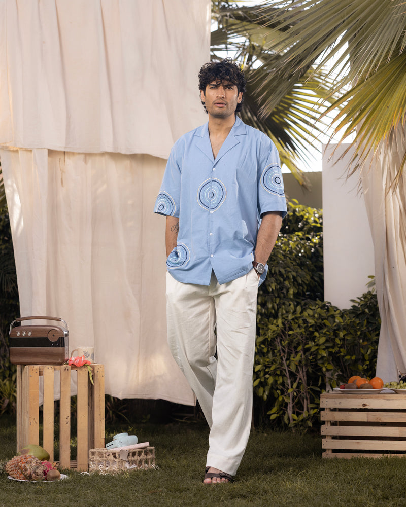 Man in a light blue shirt and white pants standing outdoors with a casual pose.