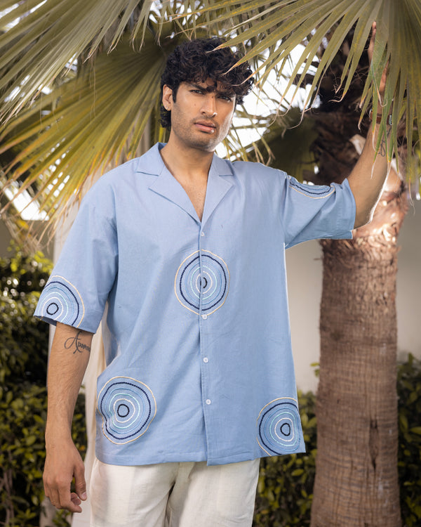 Powder blue cotton flex men’s shirt with abstract embroidered circular patterns, Cuban collar, and relaxed fit, styled as a summer resort wear outfit.
