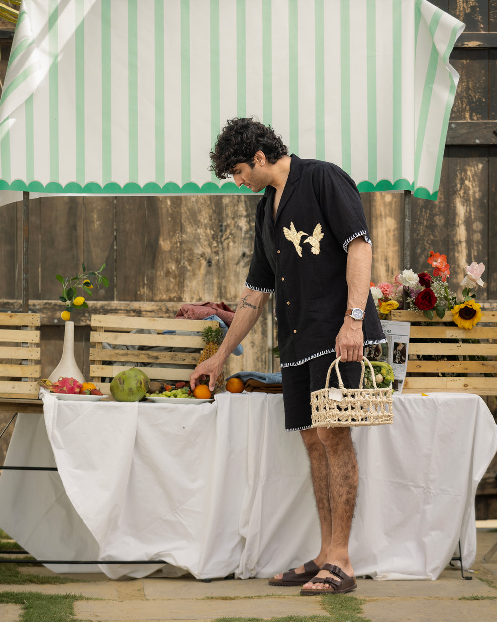 Man standing by a table with fruits and a basket, under a striped canopy.