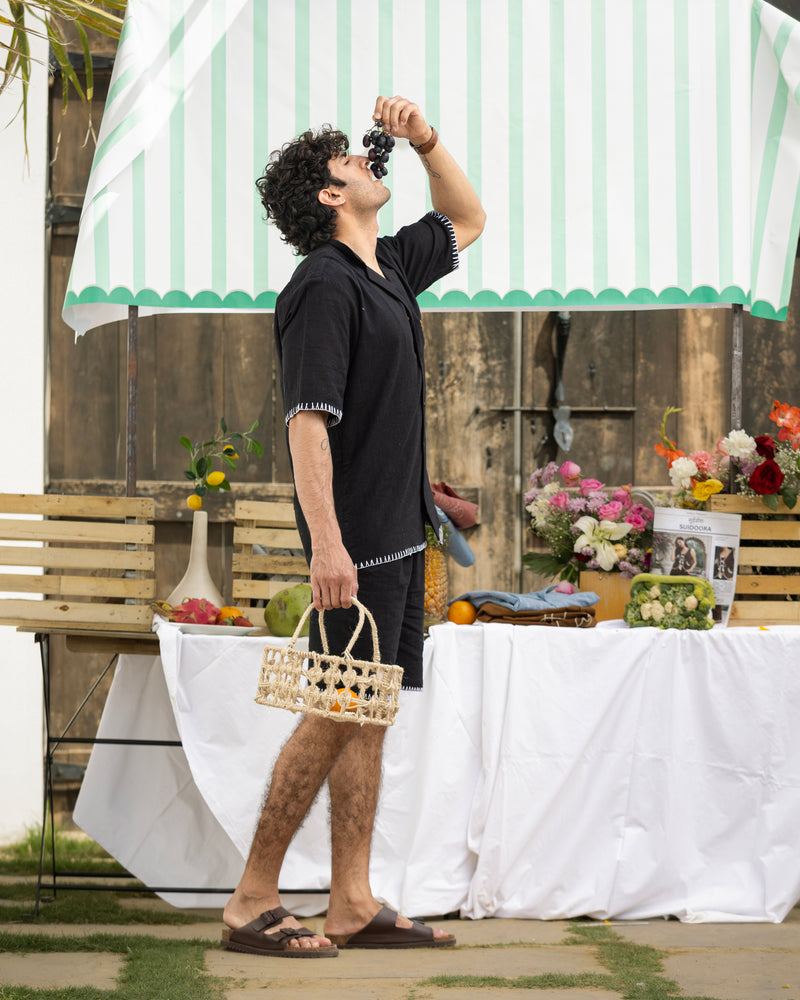 Man in black outfit holding a basket outdoors with a striped umbrella and table in the background