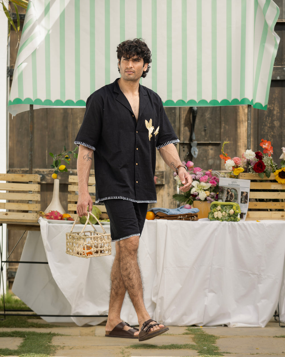 Man in black shirt and shorts standing outdoors under a striped umbrella with a table and flowers in the background.