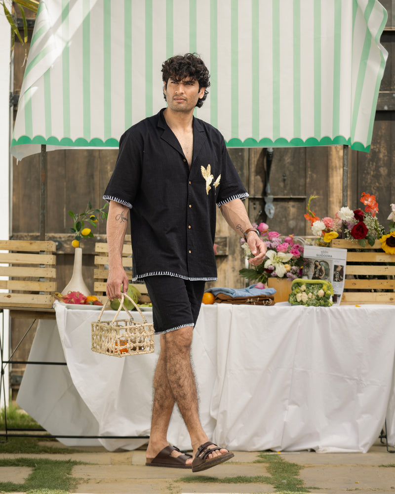 Man in black shirt and shorts standing outdoors under a striped umbrella with a table and flowers in the background.