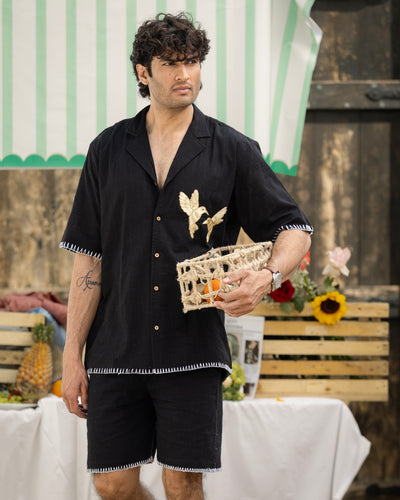 Man wearing a black outfit with floral embroidery, holding a woven basket, in an outdoor setting.
