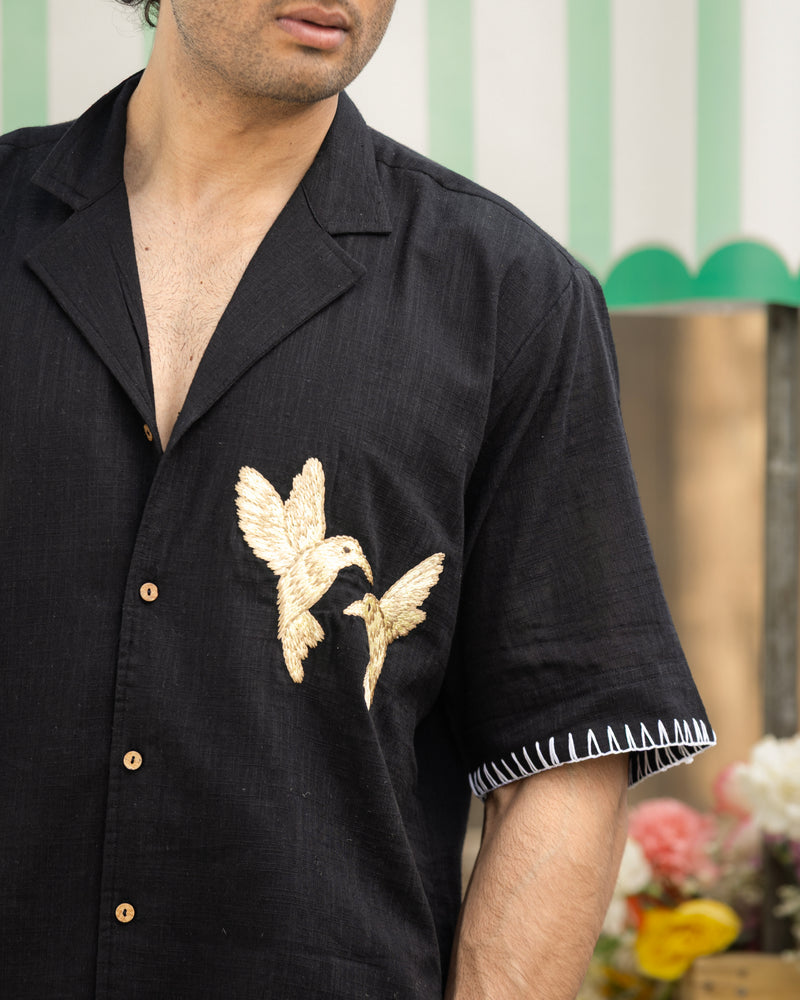 Man wearing a black shirt with gold bird embroidery, standing in front of a striped wall.