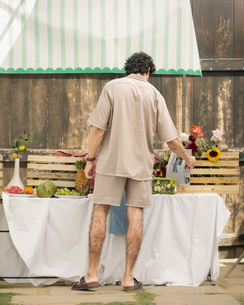 Man standing in front of a table with fruits and flowers, wearing a beige shirt and shorts.