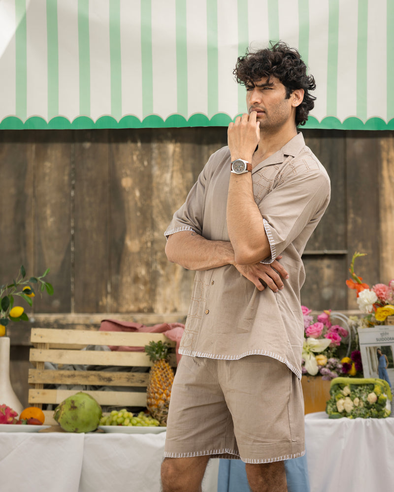 Man in beige shirt and shorts standing in front of a table with fruits and flowers, under a green and white striped awning.