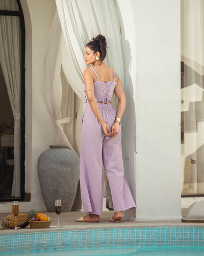 Woman in a lavender jumpsuit standing by a poolside with white curtains and decorative elements.
