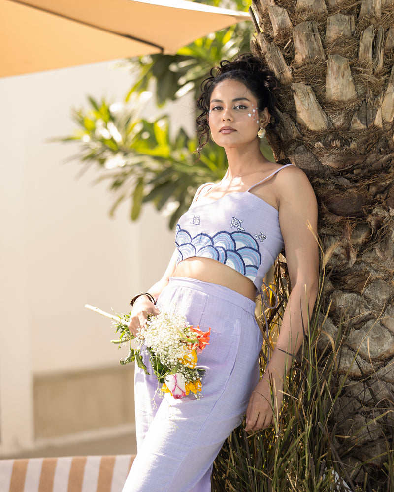 Woman in a white top and light purple pants leaning against a stone wall with plants in the background