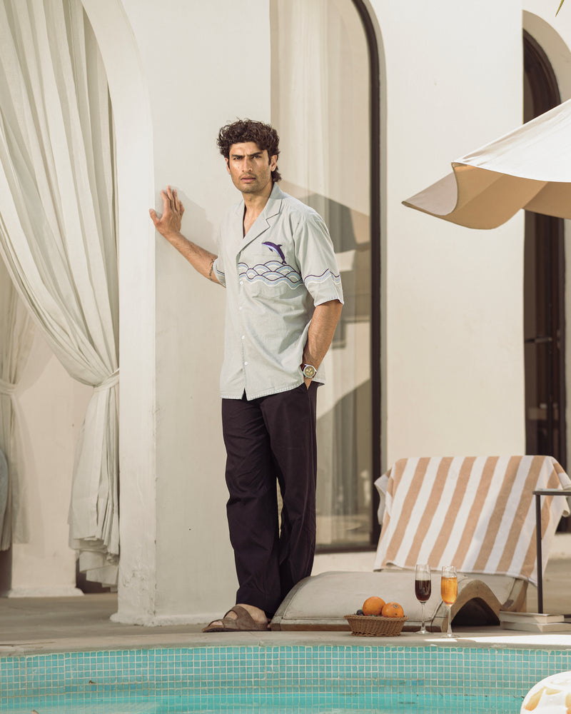 Man standing by a poolside with a white shirt and dark pants, near a striped towel and outdoor furniture.