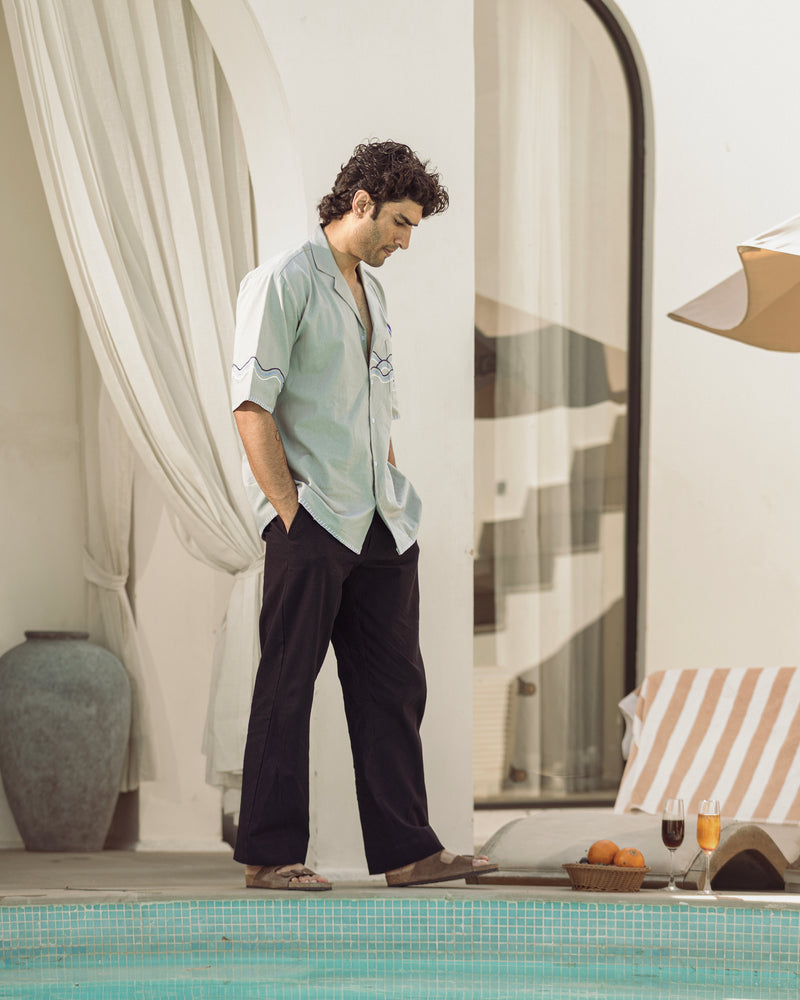 Man standing by a poolside with a relaxed posture, wearing a light shirt and dark pants.