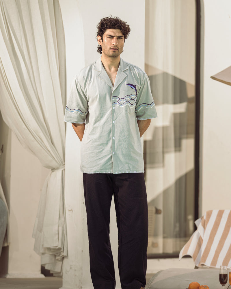 Man wearing a light green shirt with a logo and dark pants standing in a room with curtains and a chair.