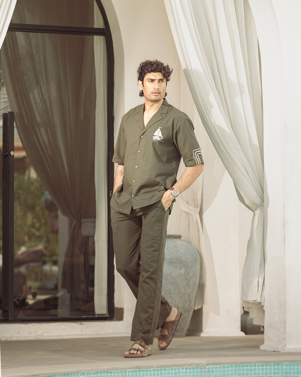 Man in green outfit standing by a poolside with white curtains in the background