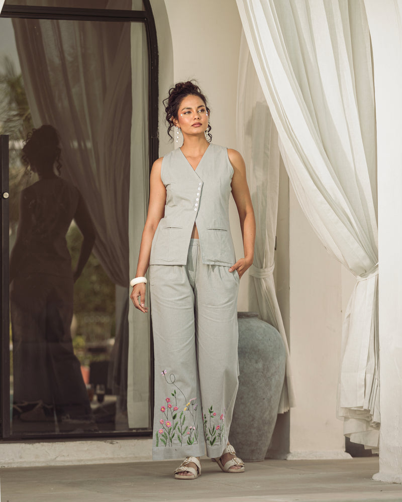 Woman in a light gray outfit with floral embroidery standing in a room with white curtains.
