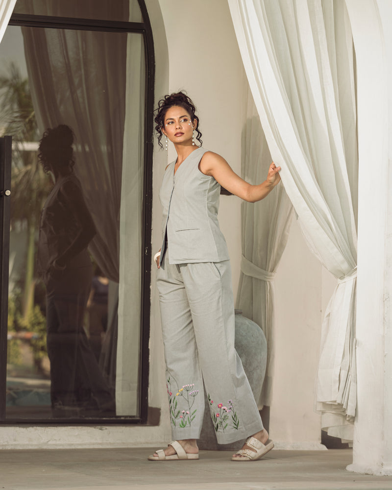 Woman in a light gray outfit with floral embroidery standing by a white curtain.