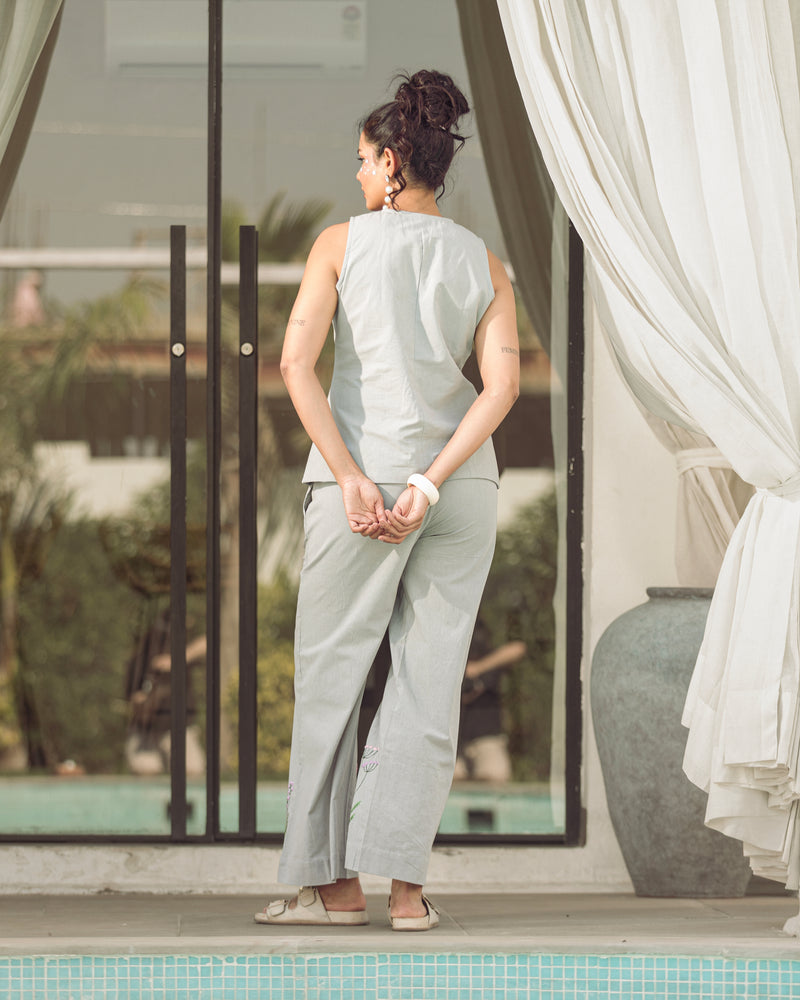 Woman in light gray sleeveless top and pants standing by a poolside with white curtains.