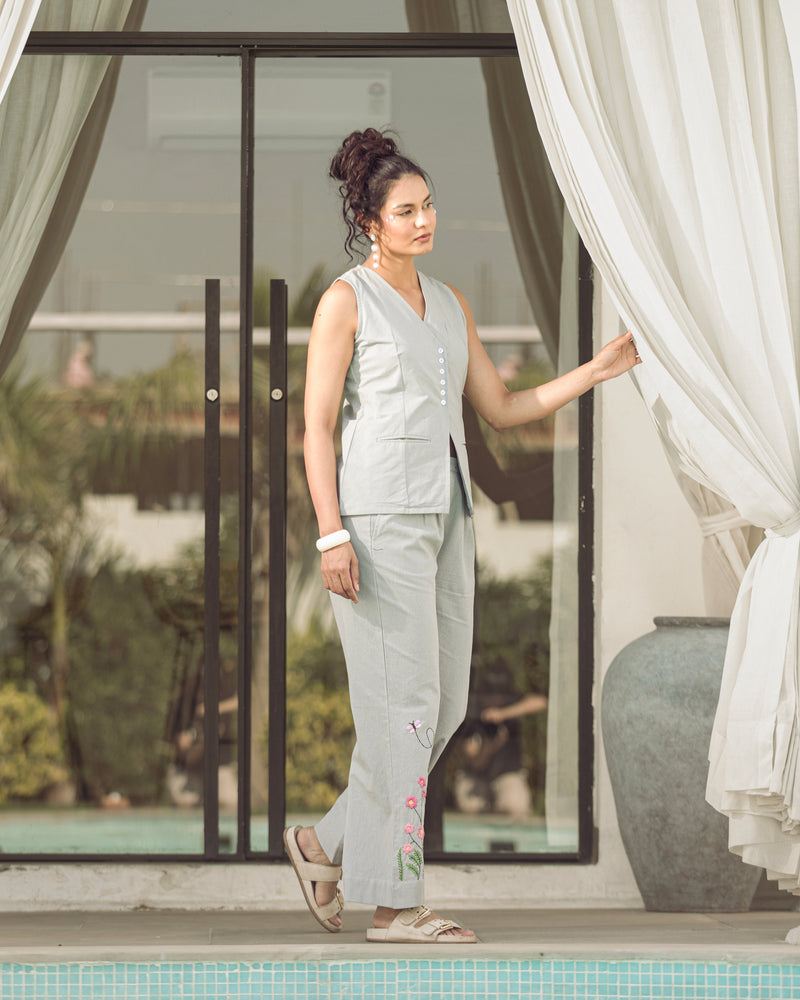 Woman in light gray sleeveless top and pants standing by a poolside with white curtains.